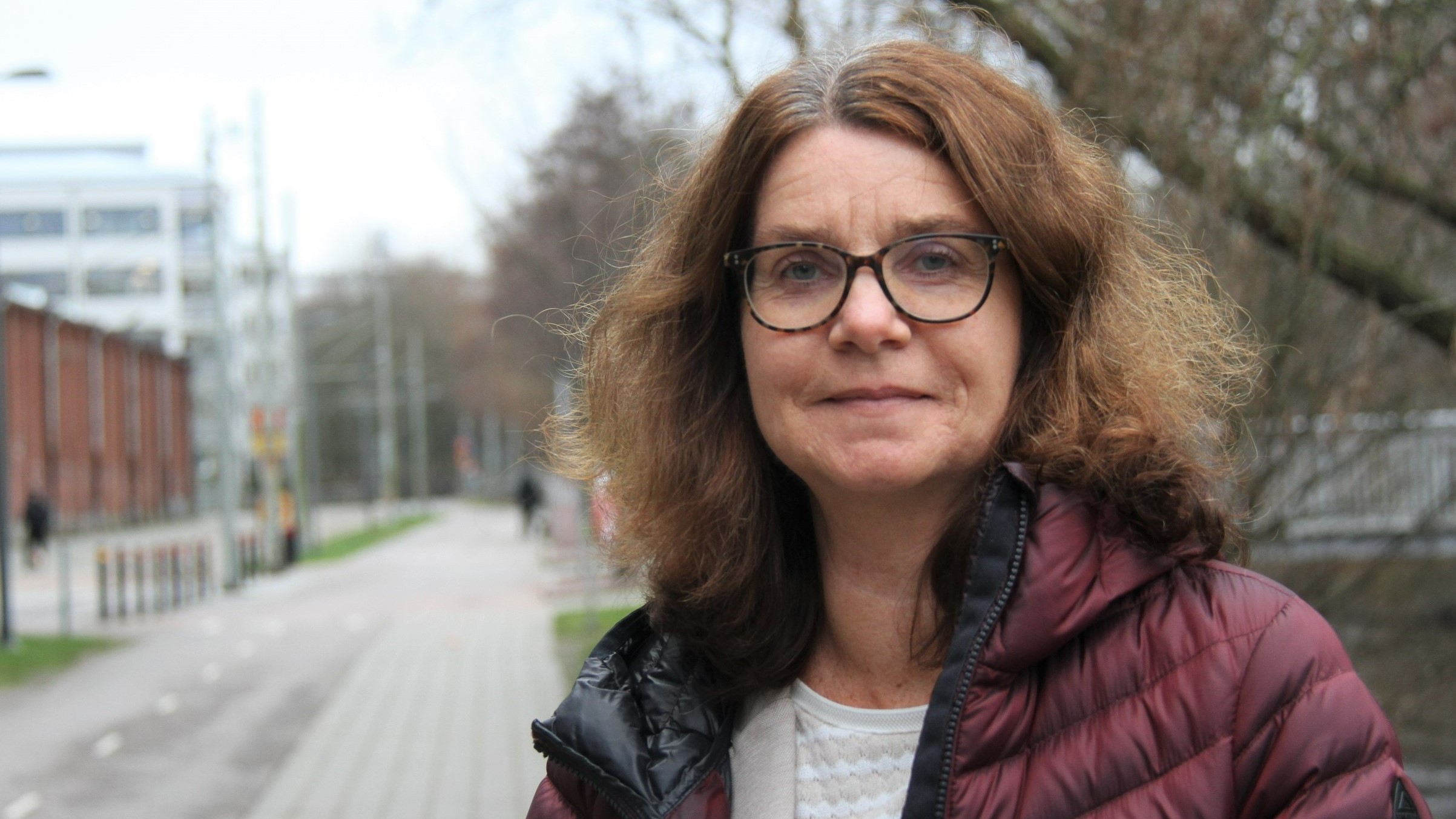 Kvinna i brunt lockigt hår som står utomhus på en gångväg i grön jacka. 
