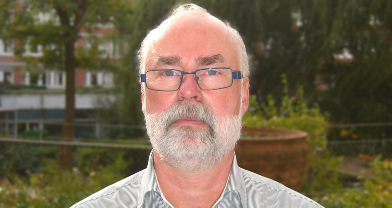 Äldre man med skägg och mustasch som står utomhus i grönska. 