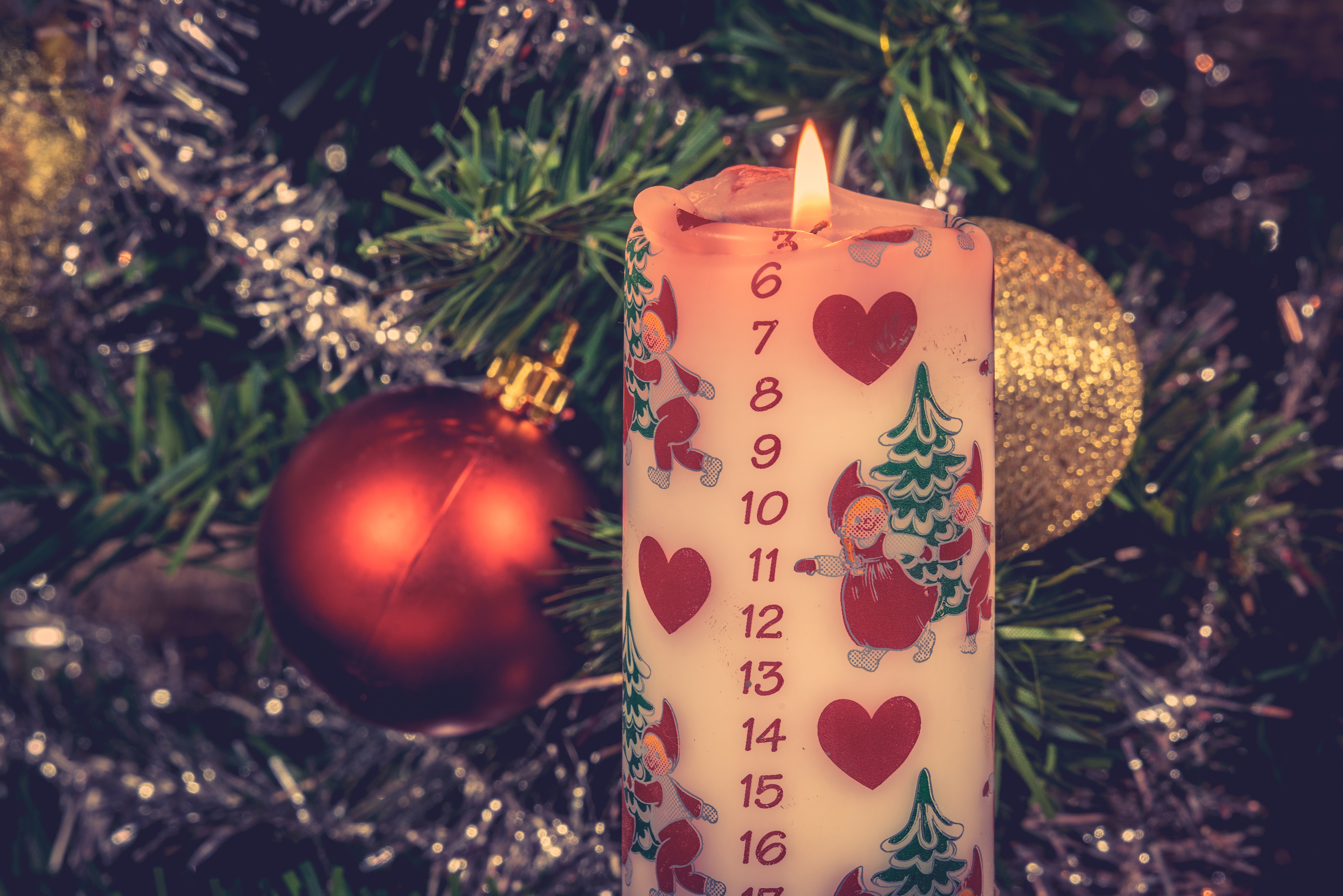 Ett kalenderljus i en plaskgran. Ljuset är tänt. Ljuset är dekorerat med tomtar. I granen hänger julgranskulor och glitter. 