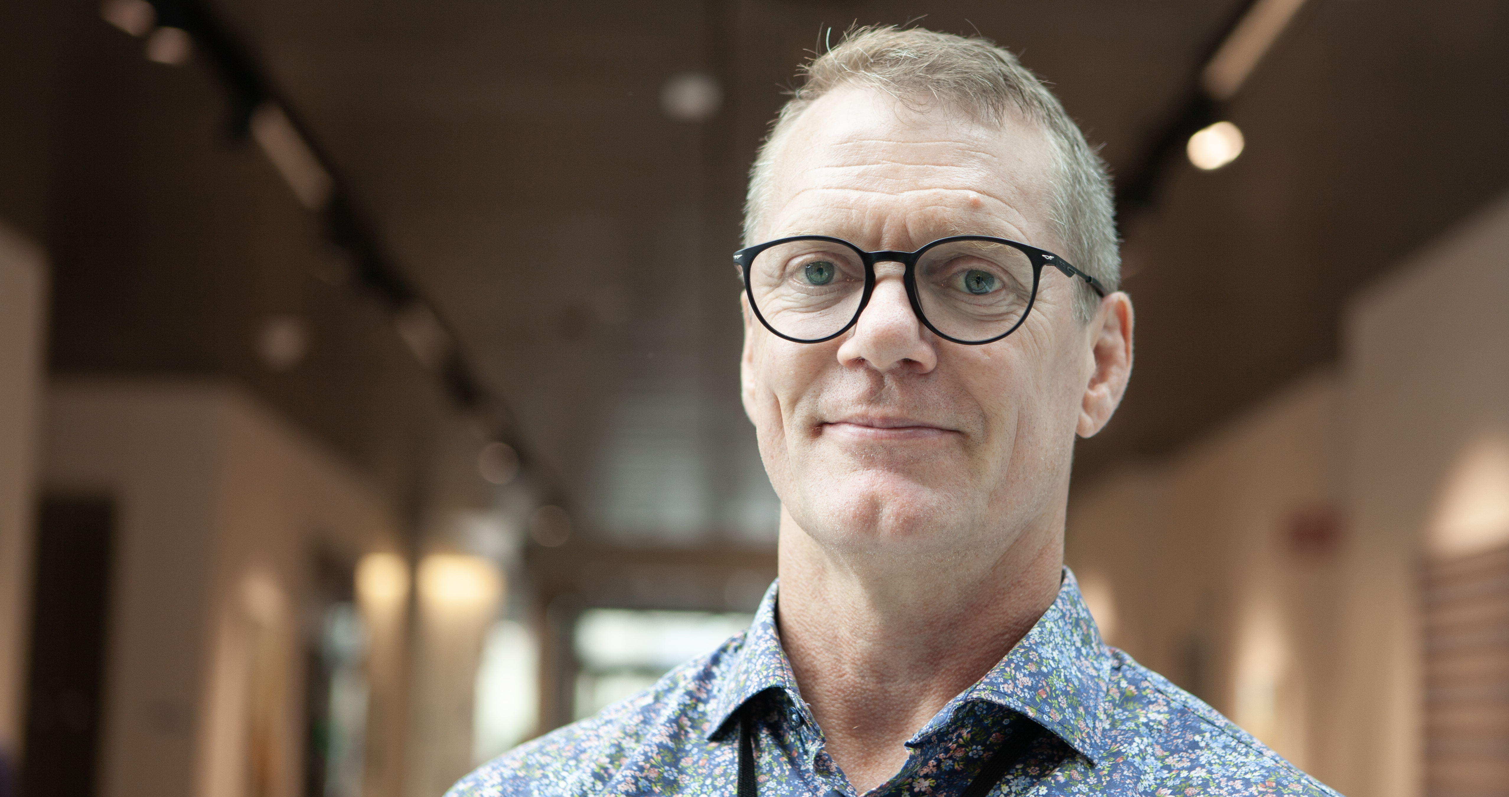 Man med kort hår, glasögon  och blommig skjorta står i kontorsmiljö