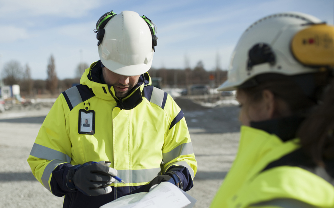 En man i varselkläder skriver på ett papper ute på en byggarbetsplats. Han har hjälm och hörselkåpor. 